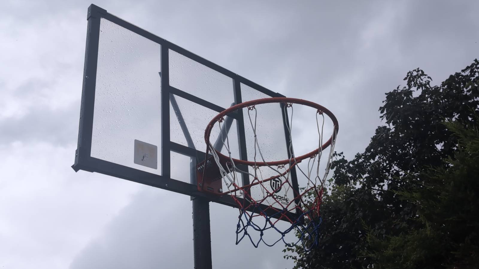 A garden basketball hoop with a clear backboard and red, white and blue net, wet with rain under a grey sky